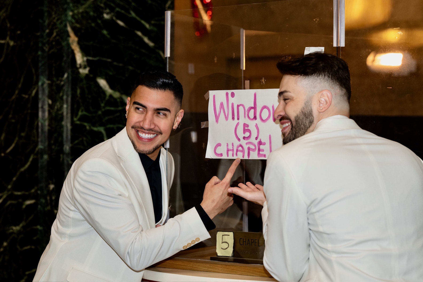 Two grooms smiling beside a City Hall chapel window sign